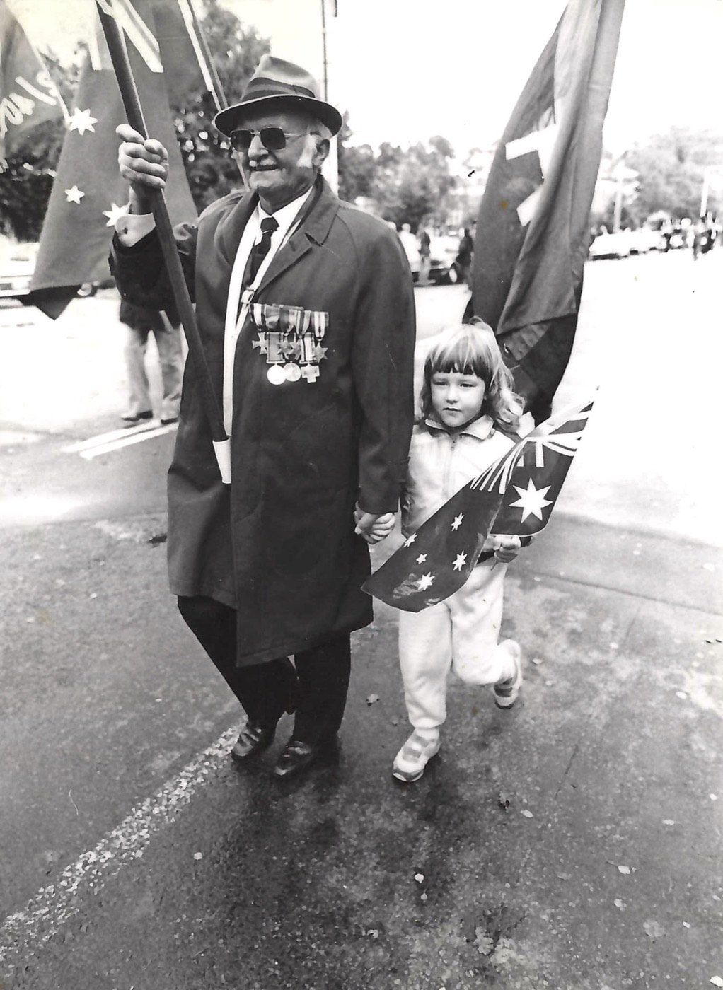 Black and white photo of Walenty (Val) in a hat and overcoat, his war medals pinned to his chest, carrying a flag and holding hands with a young Vanessa as they march on ANZAC Day